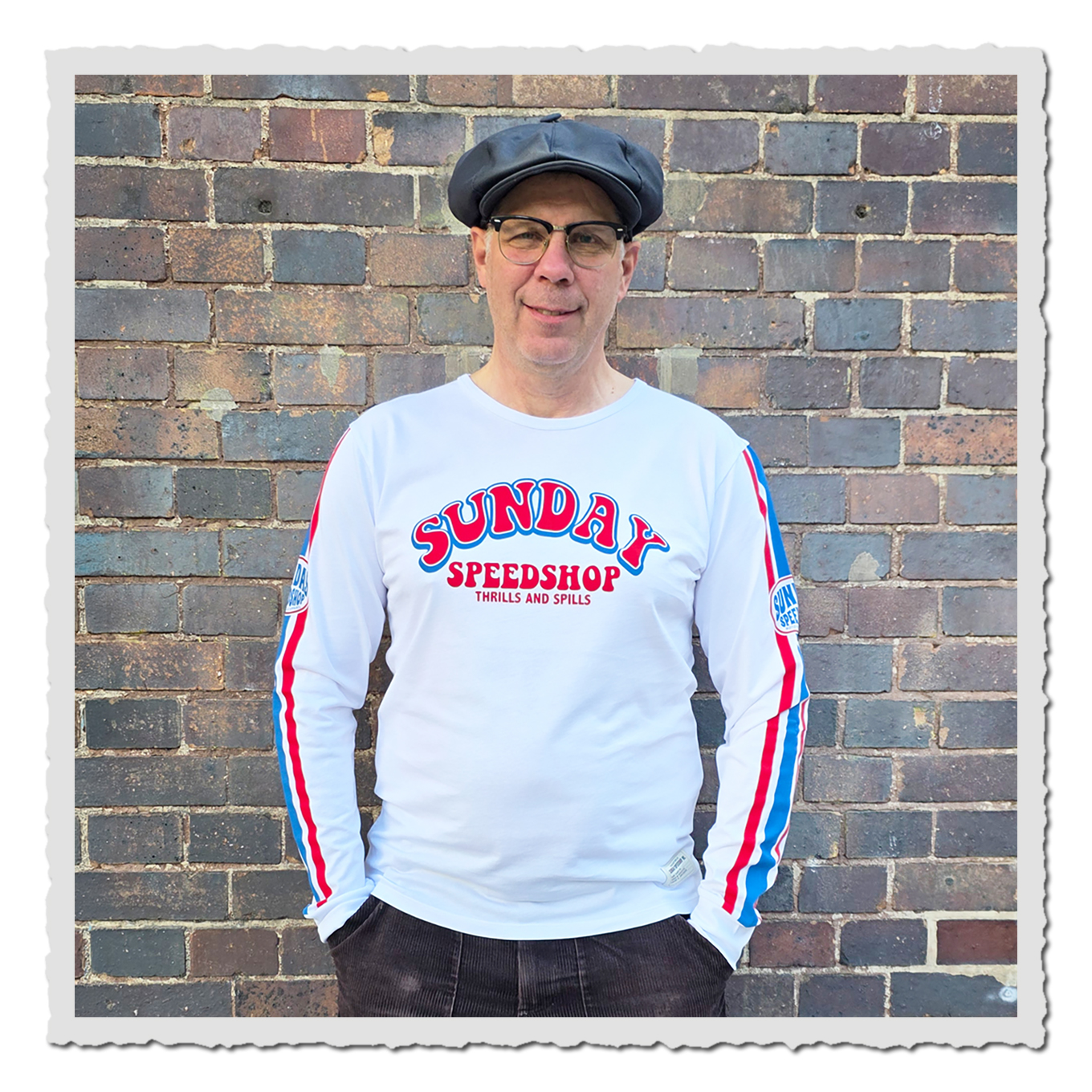 Mann trägt weißes Langarmshirt mit rot-blauem Frontdruck vor einer Backsteinwand.